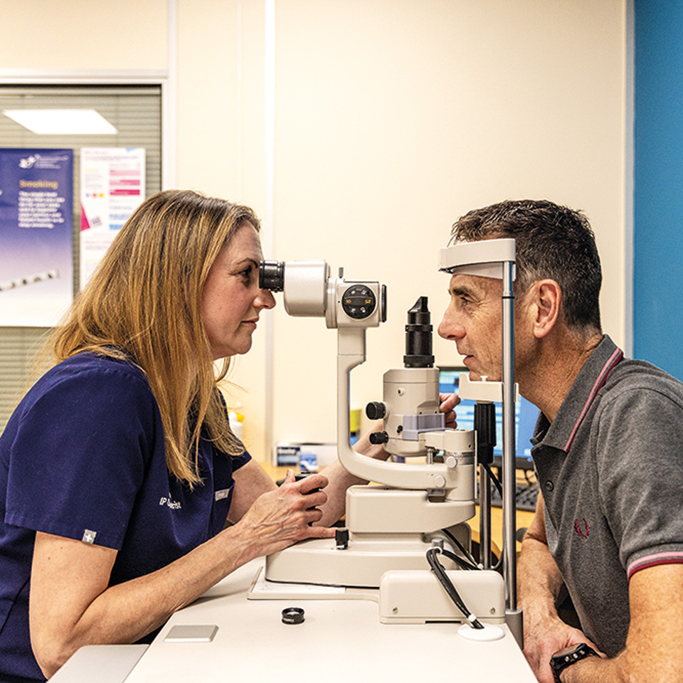 A woman with long blonde hair is photographed side on examining the eyesight of a man in a grey polo shirt. She looks into equipment that resembles a microscope. 